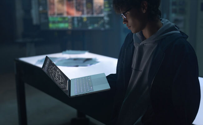 A young man in glasses reviews a 3D CAD wireframe model on a ProArt P14 laptop in a dimly lit, high-tech workshop featuring glowing screens and a light table